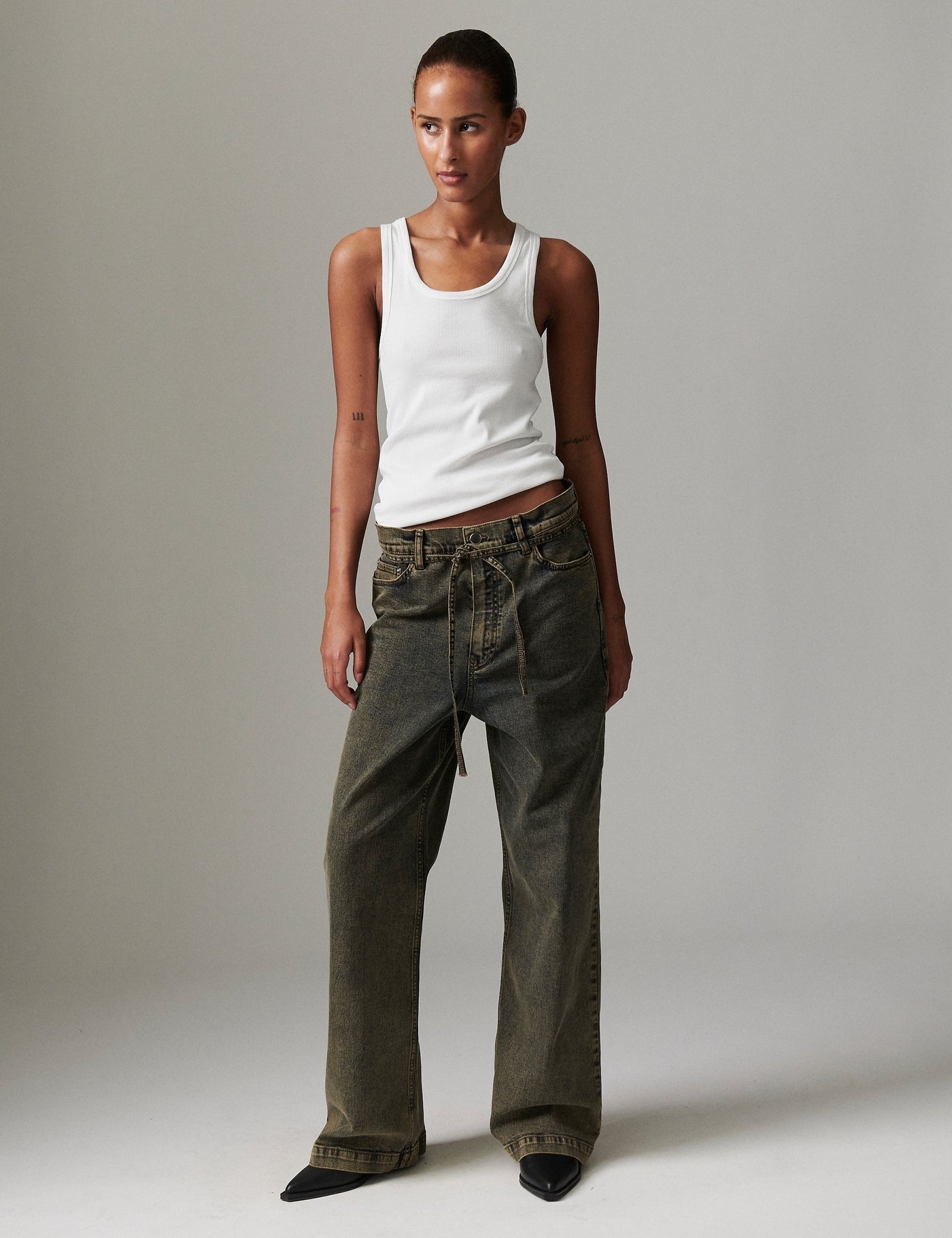 Model in DAY Birger ét Mikkelsen Elijah Elm Overdye Denim with white tank top and black pointed-toe shoes, showcasing low-rise and loose-leg silhouette.