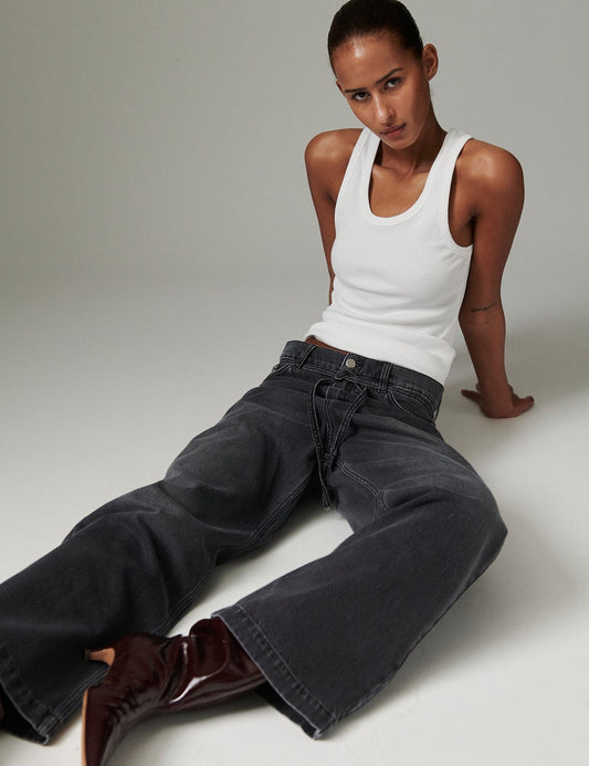 Model wearing DAY Birger ét Mikkelsen Elijah Rigid Denim in washed black, styled with a white tank top and burgundy pointed-toe boots, seated on the floor.