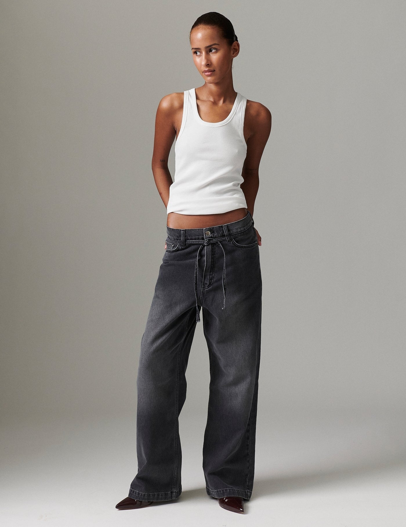 Model in DAY Birger ét Mikkelsen Elijah Rigid Denim in washed black with white tank top and burgundy pointed-toe boots, hands behind back.