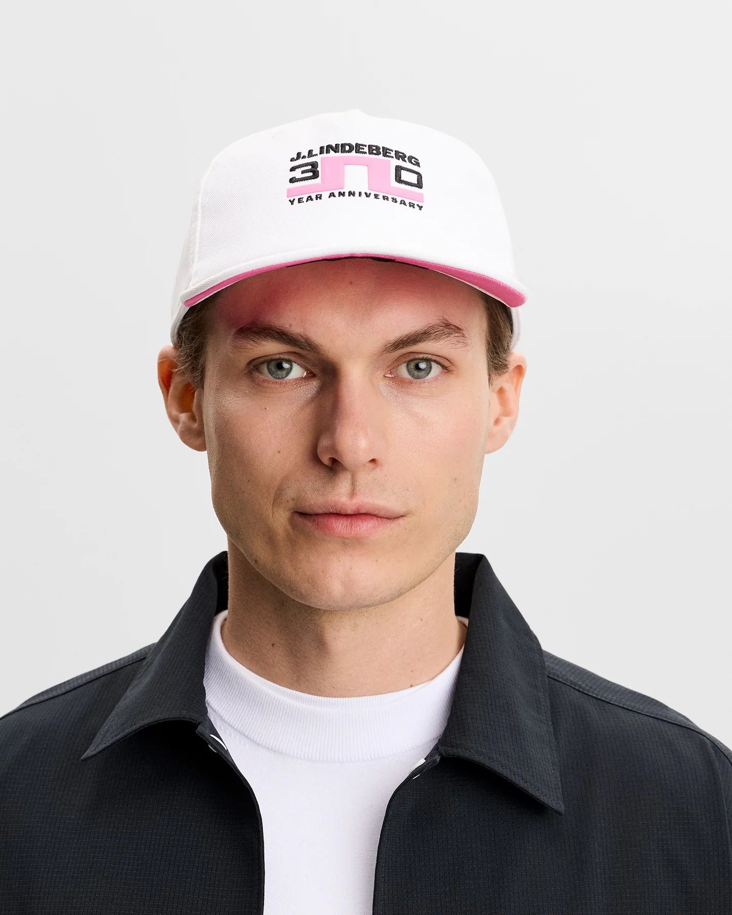 Person wearing a white cap with branding and a black jacket with embroidered text on a light gray background