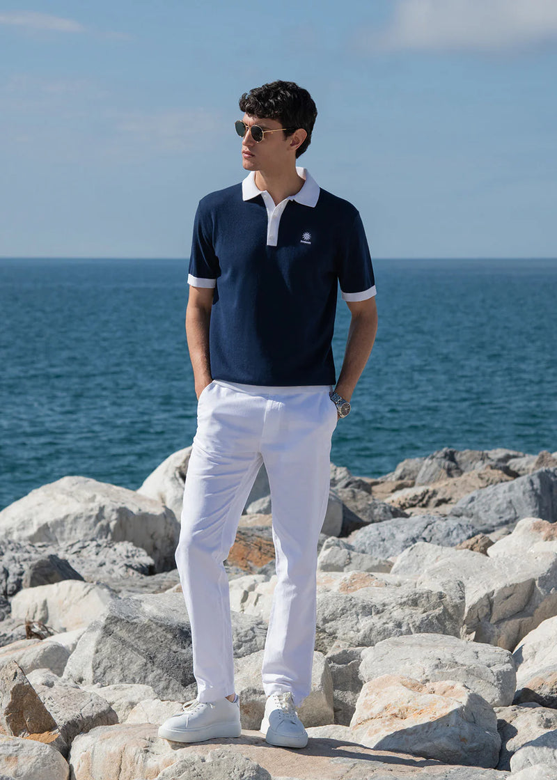 Man wearing a navy polo shirt and white pants standing on rocks with ocean in the background
