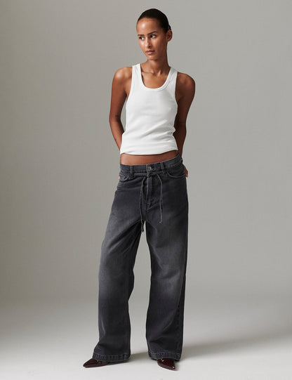 Model in DAY Birger ét Mikkelsen Elijah Rigid Denim in washed black with white tank top and burgundy pointed-toe boots, hands behind back.