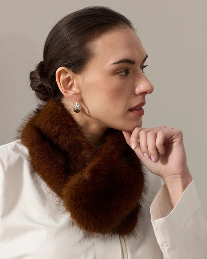 A woman in profile wearing a luxurious dark brown fur collar or cowl over a cream-colored jacket, set against a plain light gray background.
