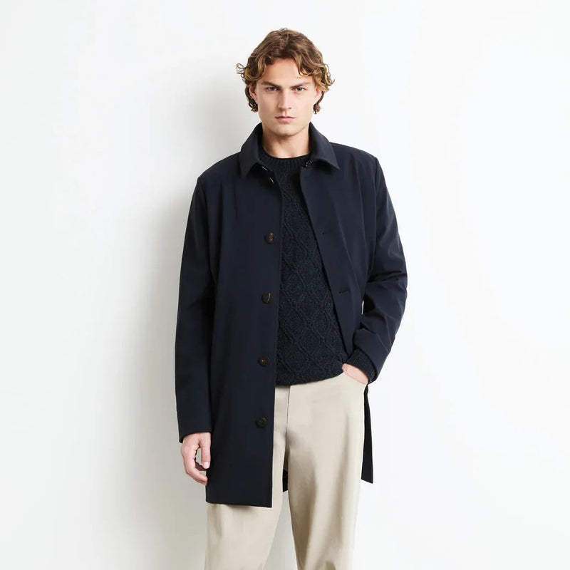 A man modeling a dark navy longline coat over a dark textured sweater, paired with beige trousers and gray sneakers, standing against a plain white wall.