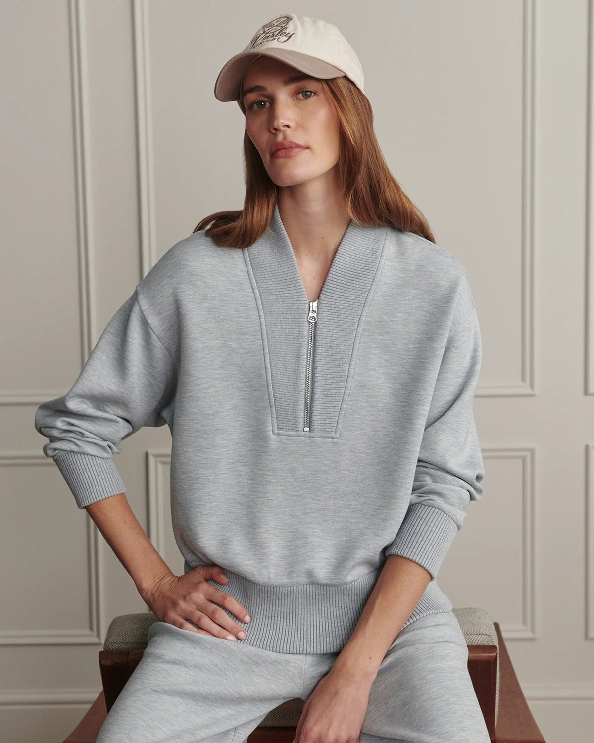 A woman wearing a light gray heathered lounge set consisting of a half-zip pullover sweater and matching sweatpants, paired with a beige baseball cap, sitting on a wooden chair against a paneled wall.