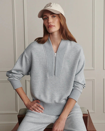 A woman wearing a light gray heathered lounge set consisting of a half-zip pullover sweater and matching sweatpants, paired with a beige baseball cap, sitting on a wooden chair against a paneled wall.
